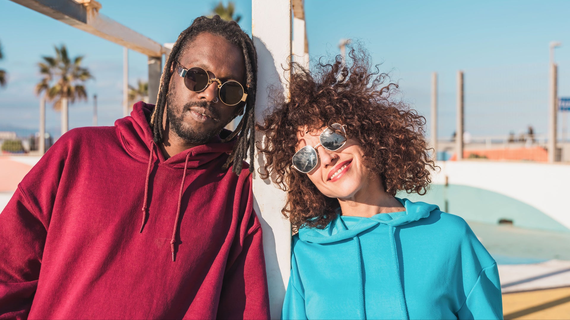 Two people wearing sunglasses and hoodies standing against a beach backdrop with palm trees.